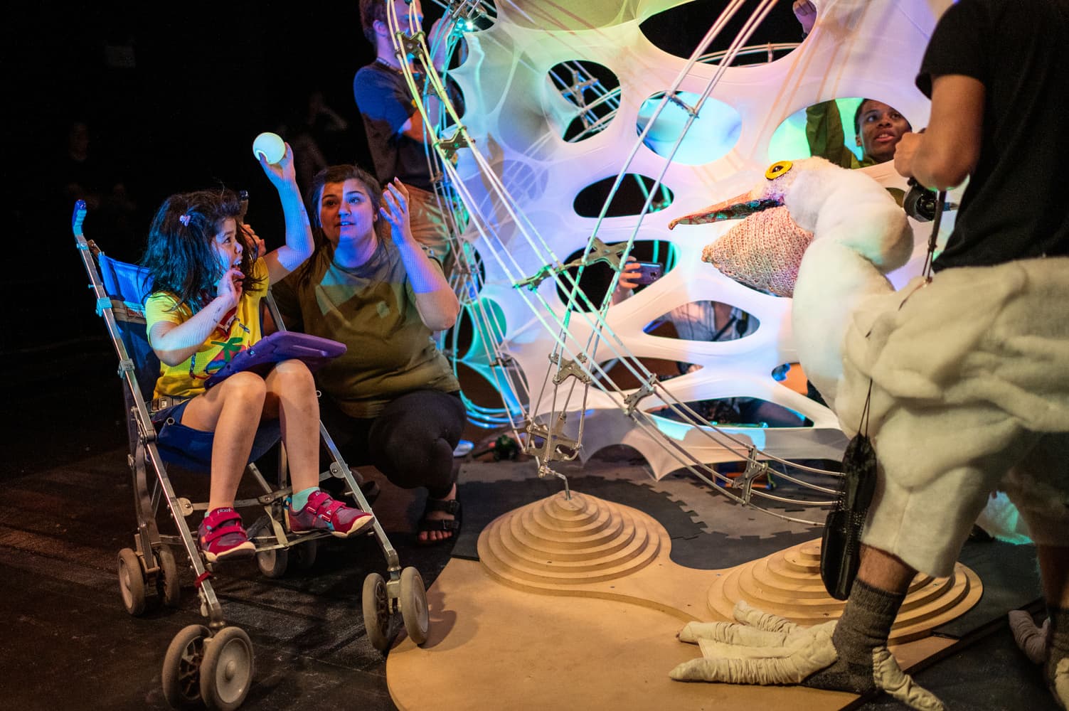 A child in a stroller and a caregiver interacting with an illuminated tensile-fabric structure and a life-sized bird puppet on stage