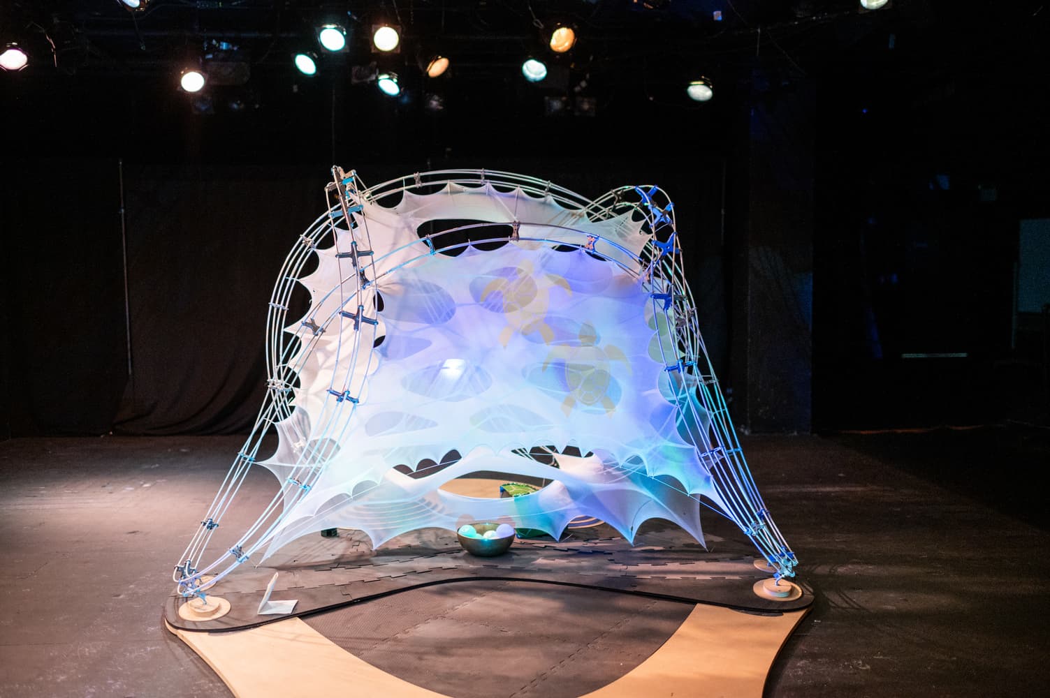 A full view of the glowing tensile-fabric pavilion, lit in blue and gold with projected turtle imagery